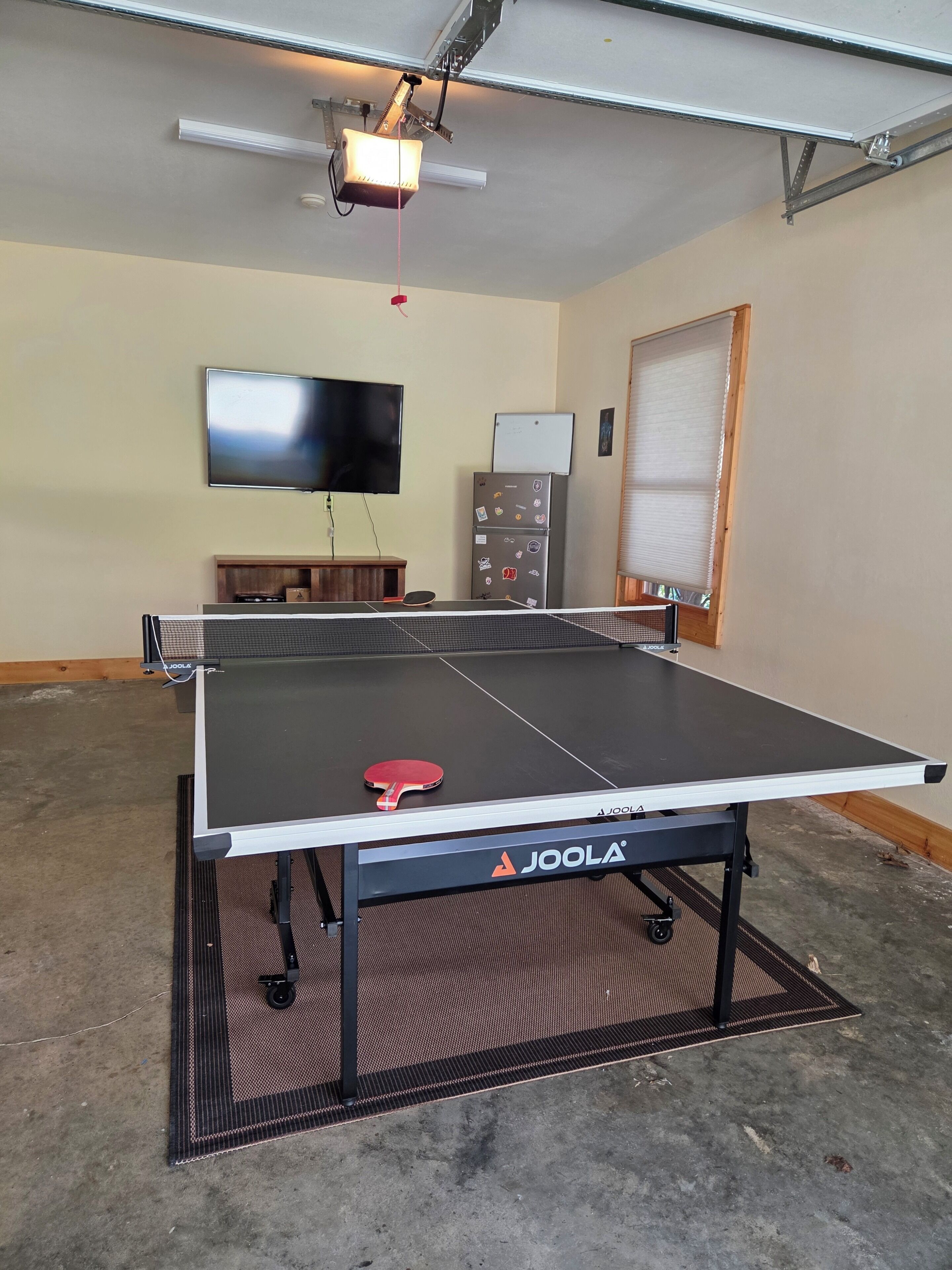Una mesa de ping pong con marca Joola, un televisor, un refrigerador y una ventana con persianas.