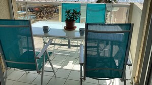 A balcony with a table and chairs, overlooking a beach with people and a sailboat.