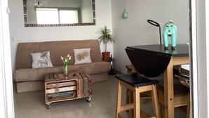 A living room with a brown sofa, a wooden coffee table, a small dining table with a black chair, a potted plant, and a mirror on the wall.