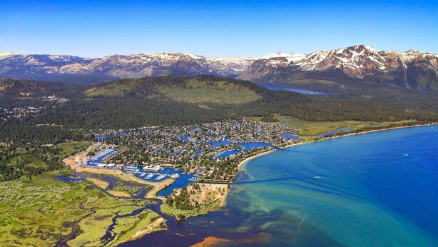 Vista aérea de un pueblo costero con un puerto deportivo, rodeado de montañas y una extensa masa de agua.
