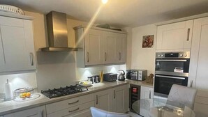 A compact kitchen with a gas stove, a toaster, a kettle, and a microwave. There is a glass table with a chair, and a small oven.