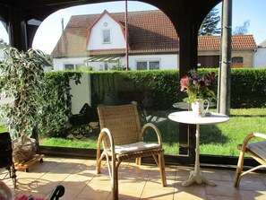 Un patio couvert aménagé avec des meubles en osier, une table ornée d’un vase de fleurs et une vue sur une maison dont le toit est recouvert de tuiles rouges.