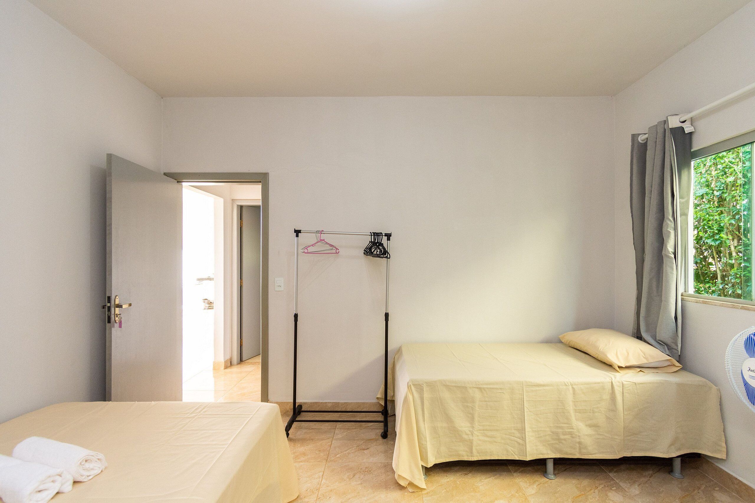 Une petite pièce propre, avec un lit, une table de chevet, un portant à vêtements et une fenêtre avec des rideaux.