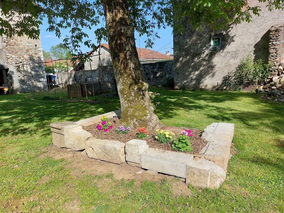 Un jardin avec un arbre et des parterres de fleurs, entouré de murs en pierre et avec un bâtiment visible au loin.