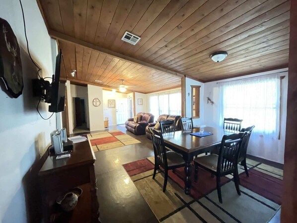 Un espacio habitable amplio con techo de madera, una mesa de comedor con sillas, un sofá y un televisor montado en la pared.