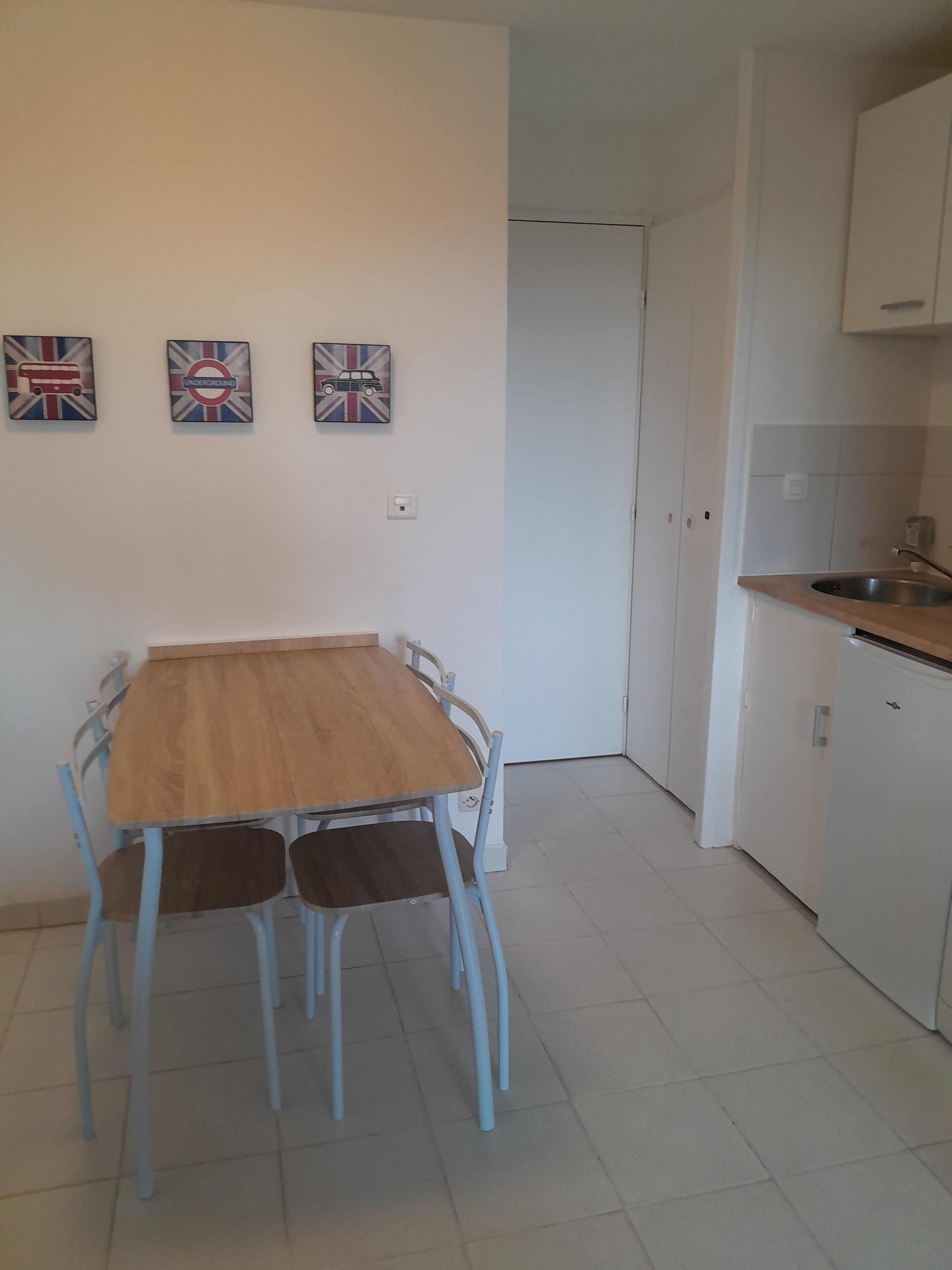 Een eetruimte met een houten tafel en blauwe stoelen, een keuken met witte kasten, en drie aan de muur hangende afbeeldingen met Britse motieven.