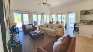A spacious living area with a sofa, a coffee table, and a dining table. There is a ceiling fan and a view of the outdoors through large windows. - Exclusive Bimini Bay home with private boat slip. (Bimini)