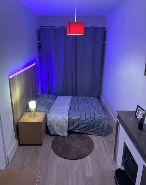 A small, narrow room with a bed, a nightstand, a red hanging light, and a fireplace.