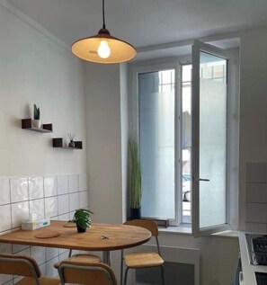 A dining area with a round table, chairs, a hanging light fixture, a potted plant, and a window with a view of the outside.