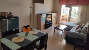 A living room with a sofa, a TV, a dining table with chairs, and a balcony with a view.