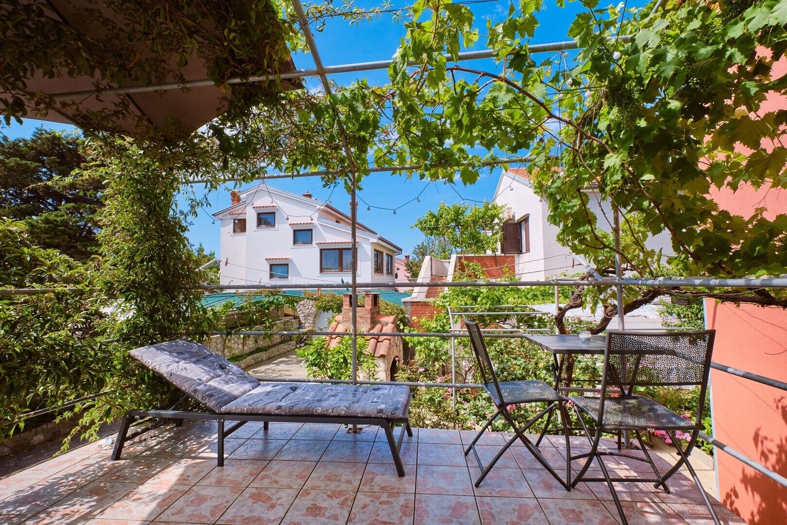 Eine Terrasse mit Tisch und Stühlen, einer Pergola und Blick auf ein weißes Haus mit rotem Dach.