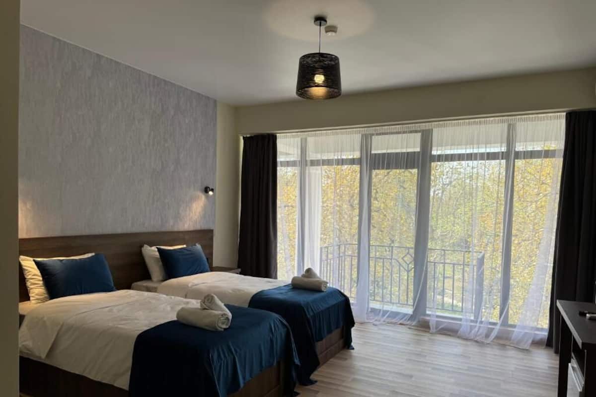 Habitación de hotel con dos camas, piso de madera, un ventanal grande con cortinas y una lámpara colgante.