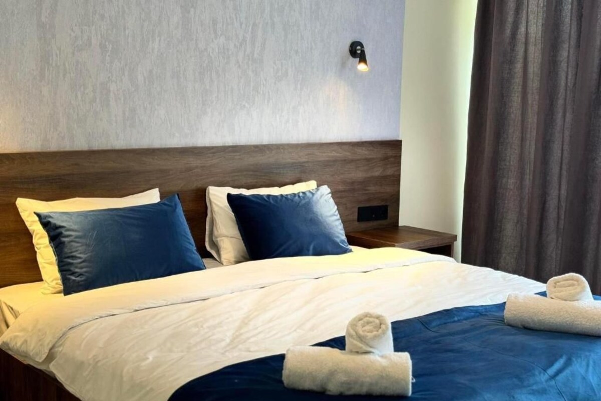 Una habitación de hotel con una cama, almohadas, una mesita de noche y una lámpara fijada a la pared.