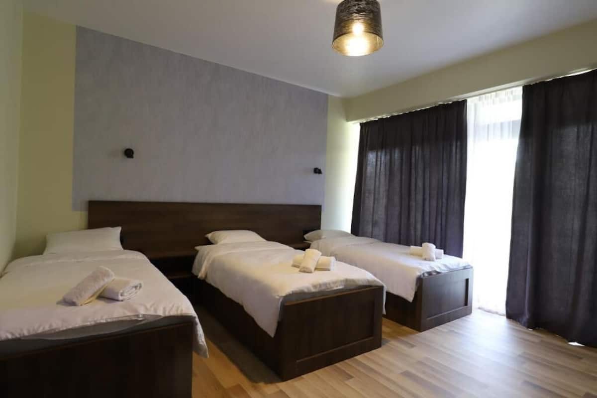 Una habitación de hotel con una cama grande, dos camas más pequeñas, piso de madera y una ventana con cortinas.