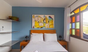 A bedroom with a bed, two bedside tables, a wall-mounted artwork, and a window with colorful glass panes. - Beautiful House in Cabo Frio Ptb0105 (Cabo Frio)