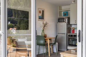 Eine moderne Küche mit Kühlschrank, Mikrowelle und Backofen. Ein Esstisch mit Stühlen und eine Vase mit Blumen stehen bereit. Ein Gemälde hängt an der Wand, und ein Fenster bietet Blick auf Bäume und Häuser.