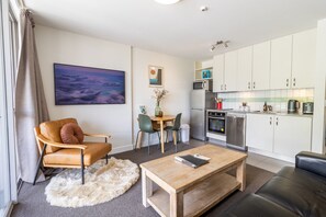 Ein modernes Wohnzimmer mit einer Couch, einem Holztisch und einem Essbereich mit Stühlen. Eine Küche mit weißen Schränken und Geräten ist sichtbar.