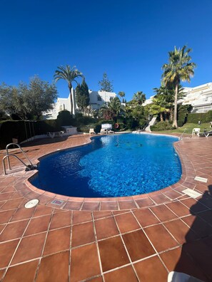 Une piscine dotée d’une terrasse carrelée, entourée de palmiers et avec un bâtiment blanc en arrière-plan.