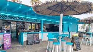 Un bar en la playa con decoración azul y blanca, mesas al aire libre y una pizarra con el menú.
