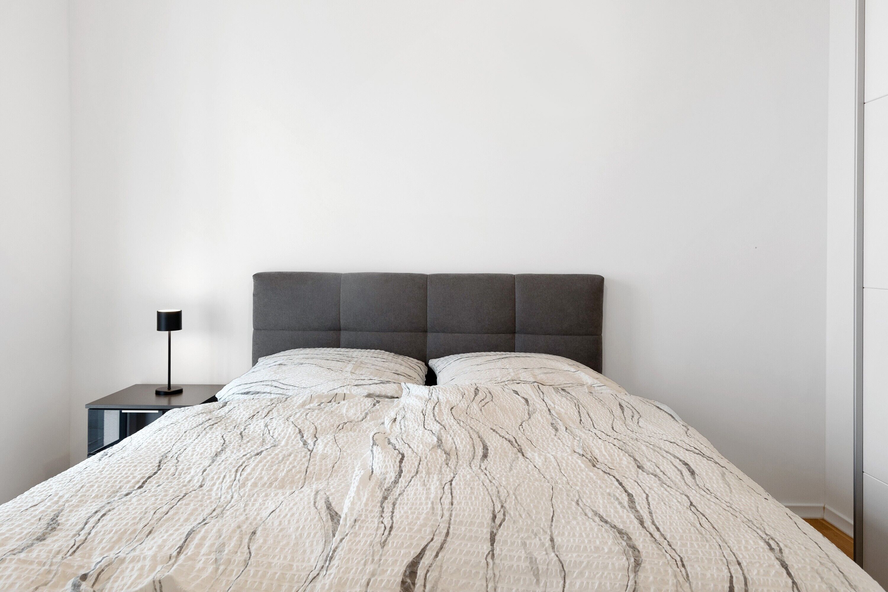 Una cama con una colcha texturizada, un cabecero oscuro y una mesita de noche pequeña con una lámpara.