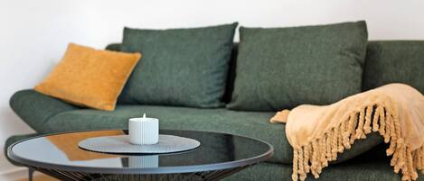 A green sofa with a mustard-colored throw blanket and a white candle on a black glass table.