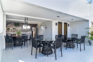An outdoor patio with a round table and chairs, a kitchen area with a brick backsplash, and a tiled wall. - Apto a 2 min da Praia em cond. c/piscina PTA0104 (Cabedelo)