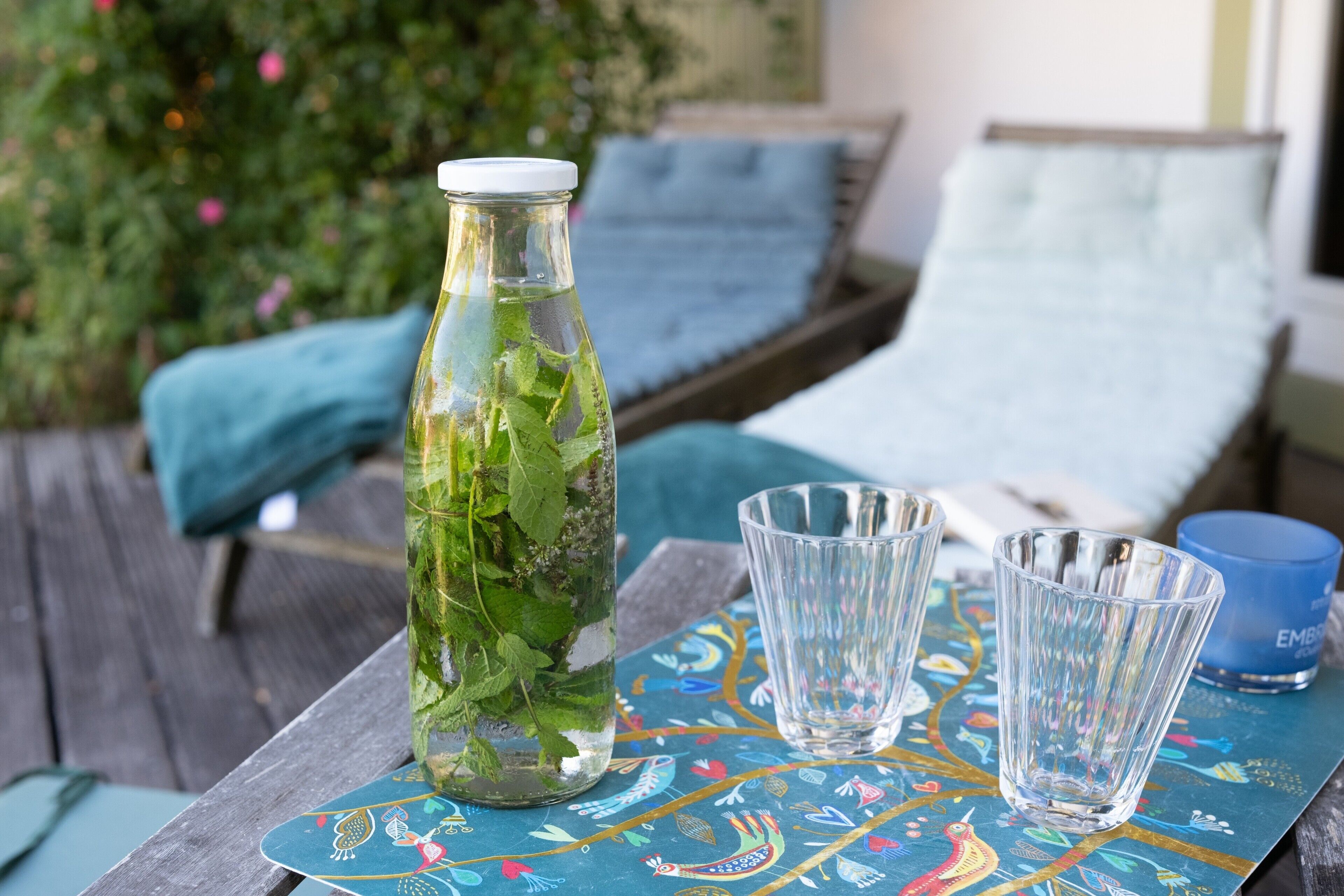 Una botella de vidrio con hojas de menta, dos vasos transparentes y una vela azul sobre una mesa de madera con mantel colorido.