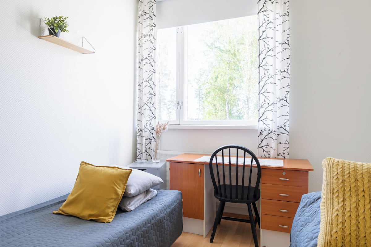 A dormitory room with a bed, desk, chair, and a window with curtains.