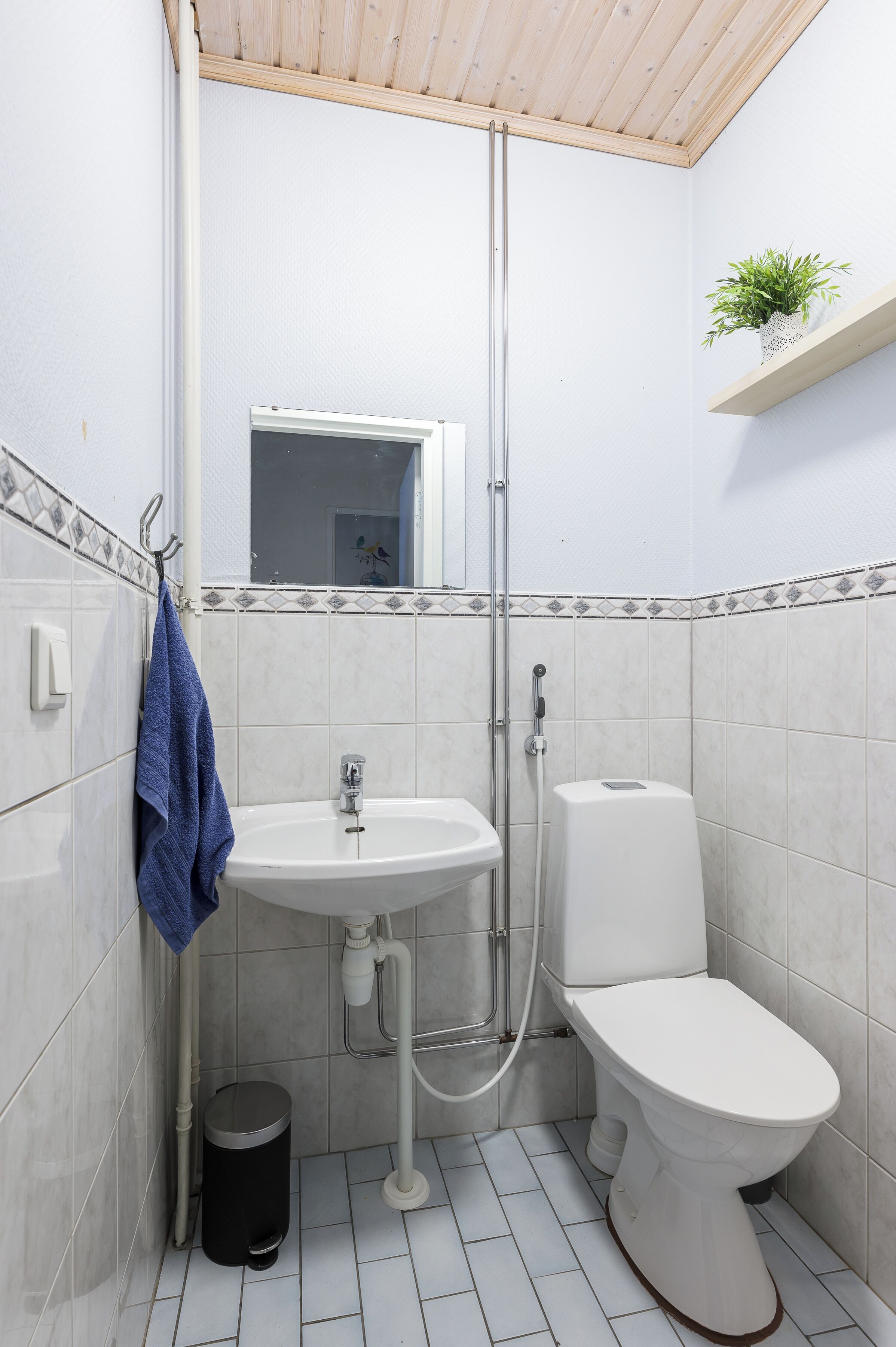 A small, clean bathroom with a white sink, toilet, and shower. There is a blue towel hanging on the wall and a small trash can.