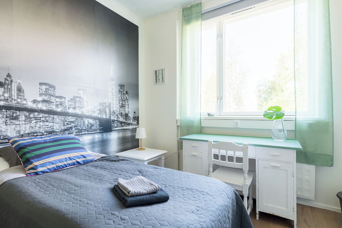 A bedroom with a bed, a desk, a window with a view, and a cityscape mural on the wall.