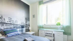 A bedroom with a bed, a desk, a window with a view, and a cityscape mural on the wall.