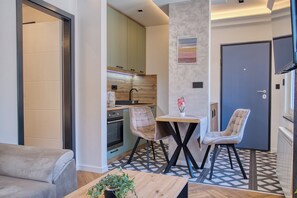 A modern living room with a sofa, a coffee table, and two chairs. There is a kitchen area with cabinets and a sink. A door is visible in the background. - Lepenička Luxe - Premium Apartments (Sarajevo)