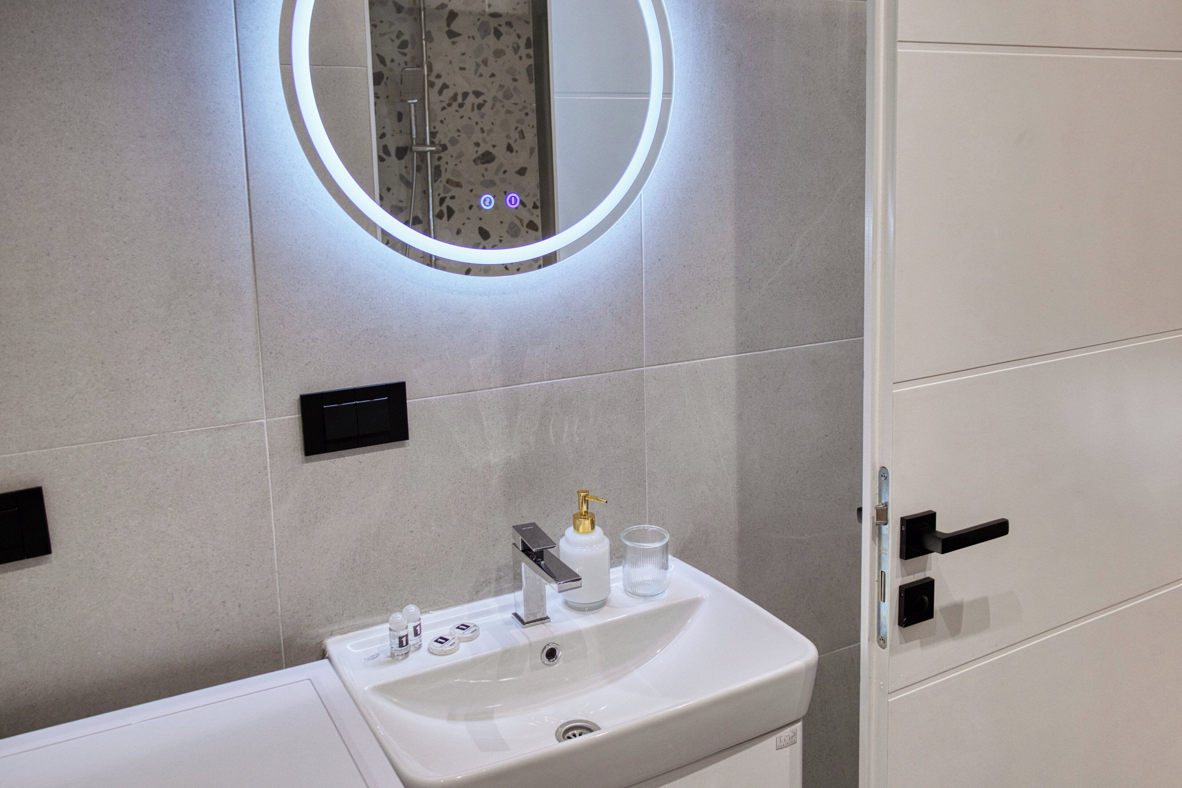 A modern bathroom with a white sink, a mirror with a circular light, and a shower area.