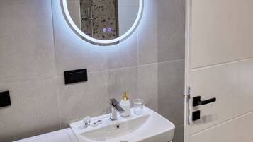 A modern bathroom with a white sink, a mirror with a circular light, and a shower area.