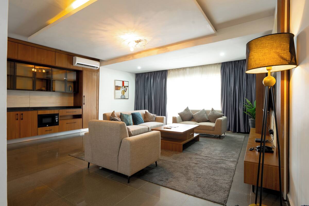 Una sala de estar moderna con un sofá, una mesa de centro y una lámpara. Cuenta con una zona de cocina con armarios y un equipo de aire acondicionado.