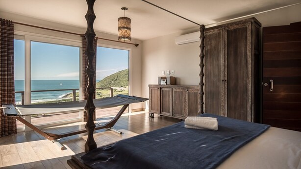 Une chambre avec une grande fenêtre donnant sur l’océan, un lit en bois et une petite table de chevet avec une lampe.