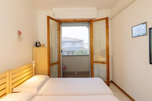 Quarto com cama, cabeceira de madeira, mesa de cabeceira, pequena mesa com abajur, janela com vista para edifícios e relógio fixado na parede.