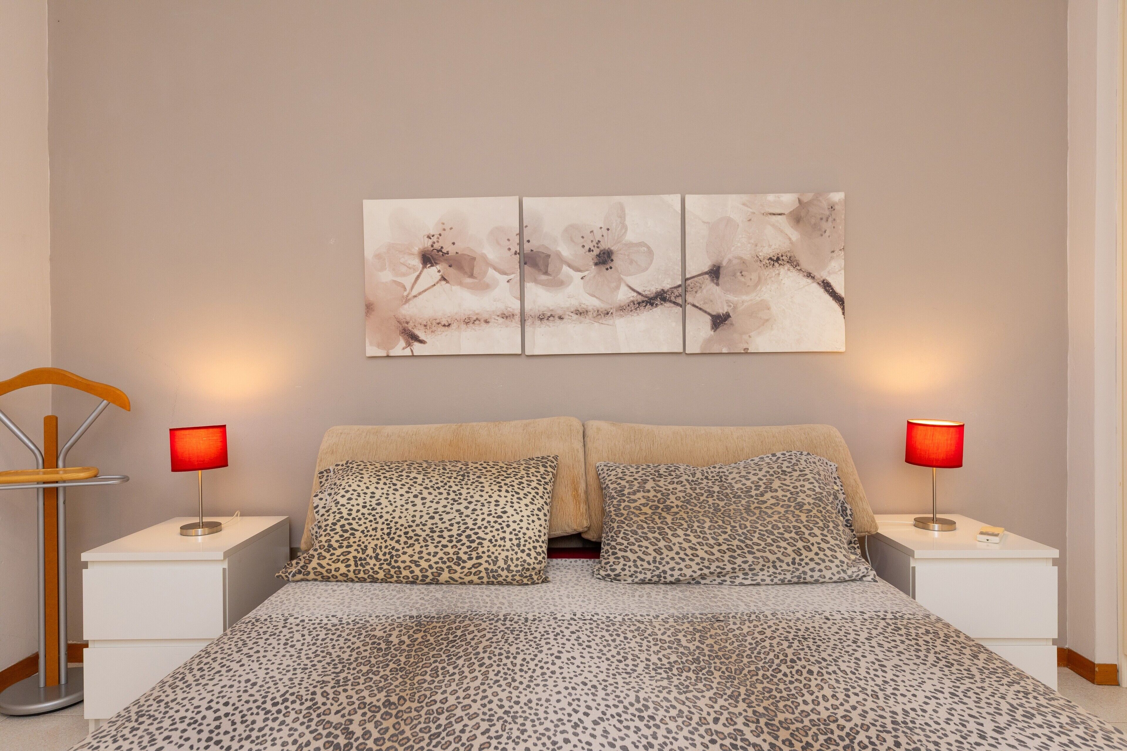 Ein Schlafzimmer mit einem Bett, zwei Nachttischen, jeweils eine rote Lampe darauf, einer Stehlampe aus Holz und einem Wandbild.