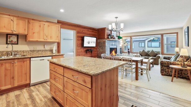A modern kitchen with wooden cabinets, granite countertops, and a built-in island. There is a dining area with a table and chairs, and a living room with a sofa and a fireplace.