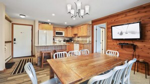 A modern kitchen with wooden cabinets, a dining table with chairs, a TV mounted on the wall, and a wooden floor.