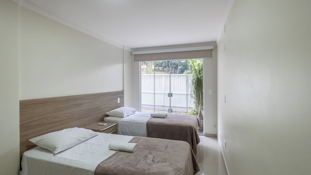 A hotel room with two beds, a wooden headboard, a large window, and a view of a fence and greenery outside. - 3BR Apt w/ BBQ 290m From Beach Pic0125 (Bombinhas)