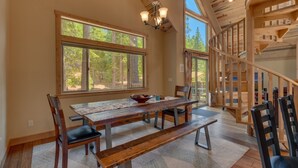 A spacious dining area with a large wooden table, benches, and chairs. There are large windows providing natural light and views of the surrounding forest. A wooden staircase is visible in the background. - The Westlands Lodge 1135ac 4 Bedroom Home (South Lake Tahoe)