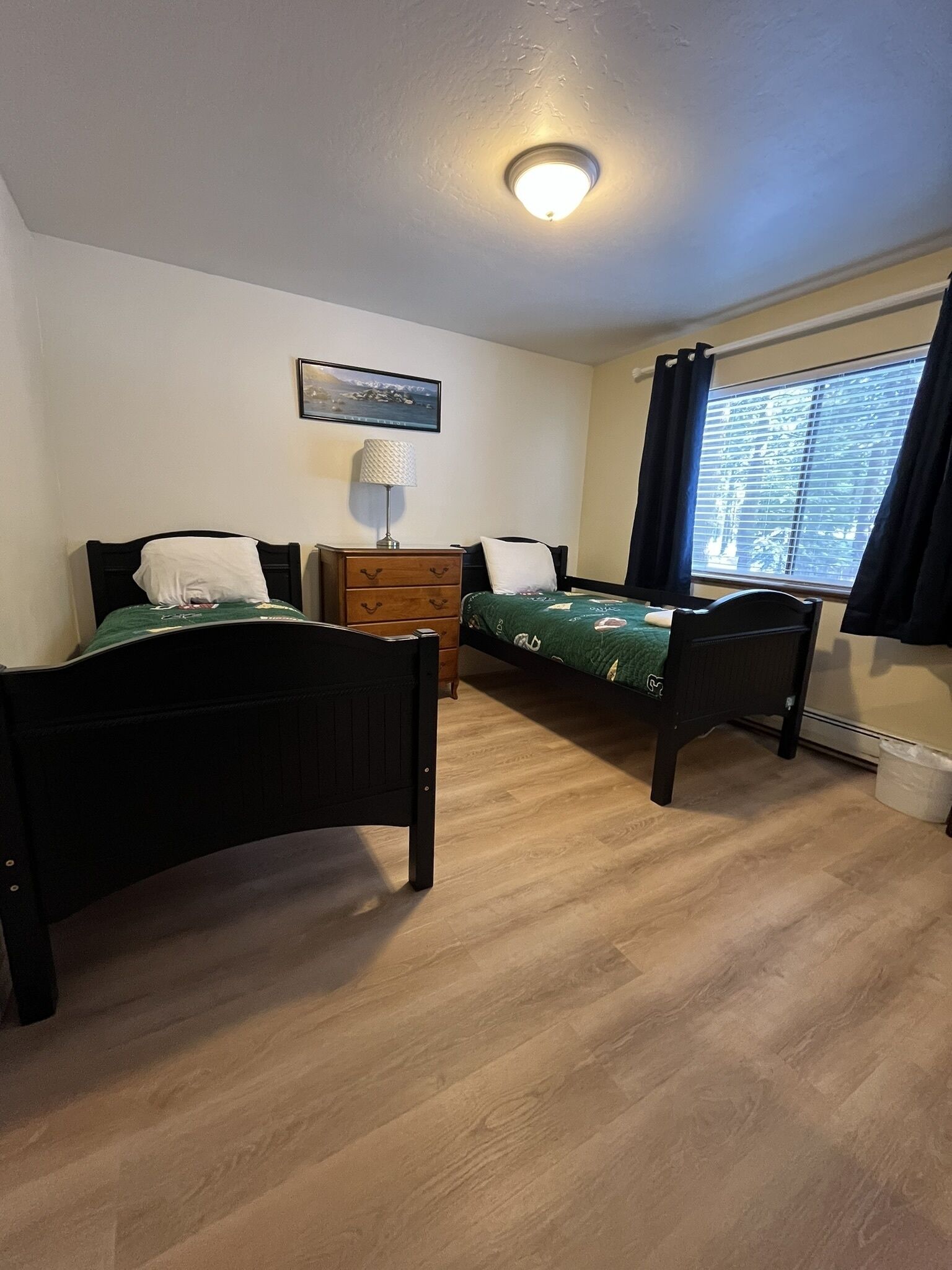 A room with two single beds, a wooden dresser, a lamp, and a window with curtains.
