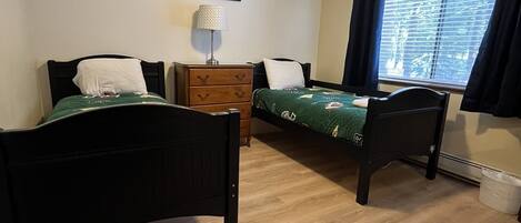A room with two single beds, a wooden dresser, a lamp, and a window with curtains.