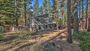 A two-story cabin with a deck, surrounded by tall pine trees and a dirt path leading to the entrance. - 121m The Meadow Drive Home 5 Bedroom Home (Stateline)