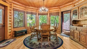 A log cabin interior with a round dining table, wooden chairs, a chandelier, and large windows offering a view of the forest. - Nature's Delight, Private Family-Friendly Cabin (McCall)
