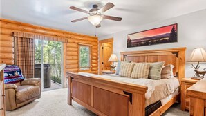 A log cabin bedroom with a wooden bed, a ceiling fan, a nightstand, a lamp, a chair, and a view of the outdoors through large windows. - Nature's Delight, Private Family-Friendly Cabin (McCall)
