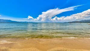 A beach with clear water, a sandy shore, and distant mountains under a blue sky.