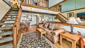 A spacious living area with a high ceiling, a wooden staircase, a dining area, and a kitchen in the background.