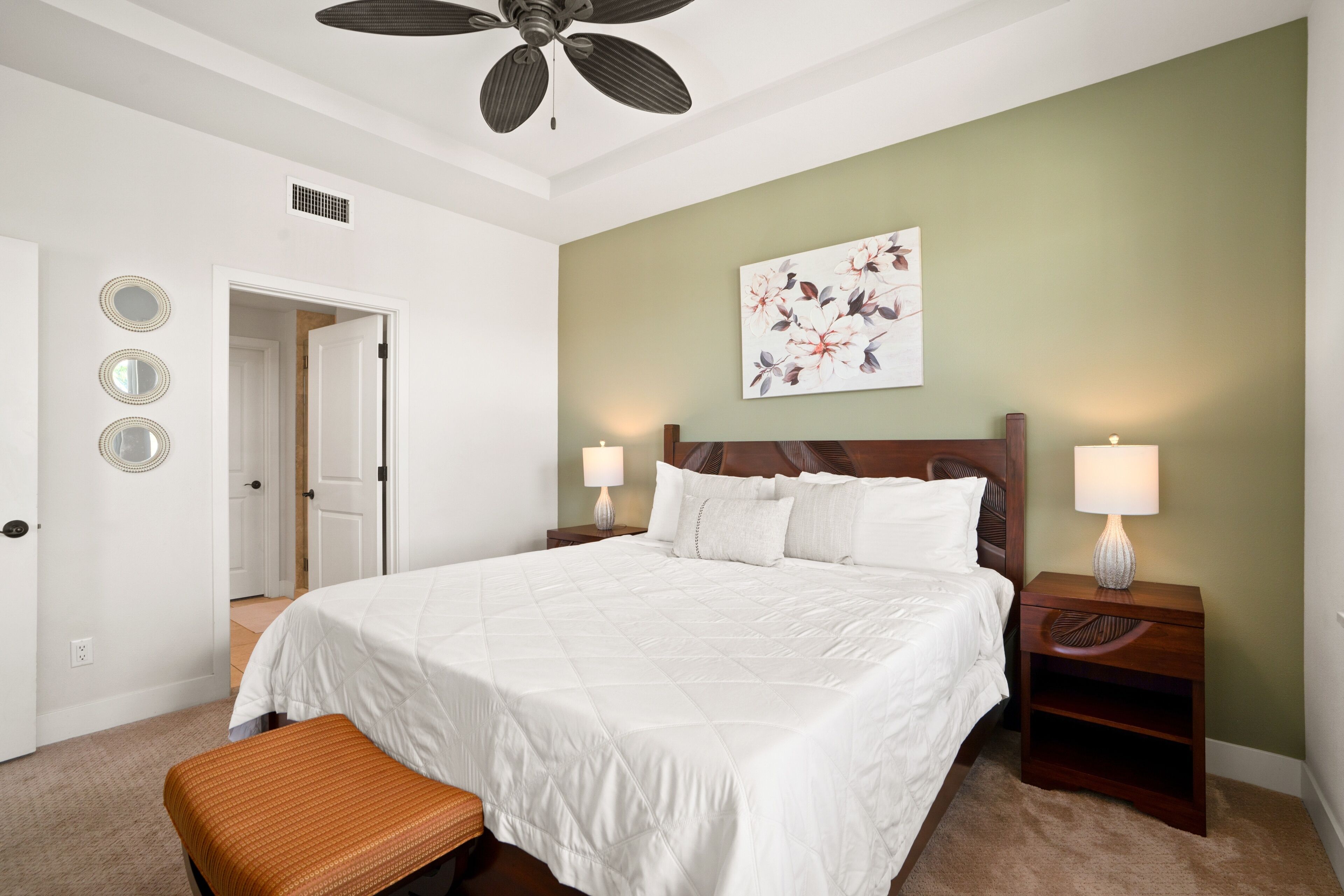 A bedroom with a large bed, two bedside tables with lamps, a ceiling fan, a door, and a wall with three circular mirrors.